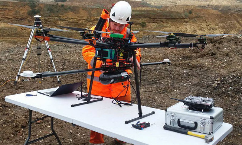 Male professional UAV pilot in orange hi-vis clothing setting up Routescene LidarPod underneath UAV in field.