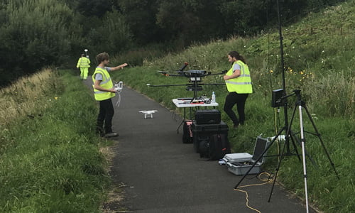 Three male surveyors in yellow hi-vis vests setting up Routescene UAV LiDAR system on a drone.