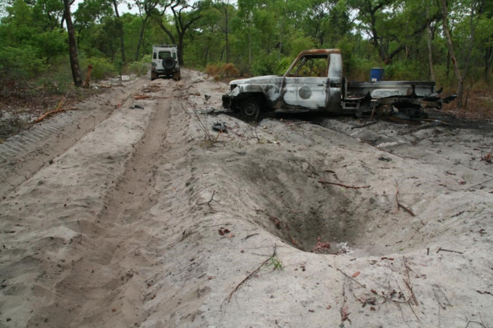 Image of sandy cleared narrow road used by the survey team to set up their equipment, with exploded mine crater and damaged truck at the side of the road.